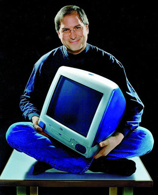 Steve Jobs holding a computer while seated on a table.